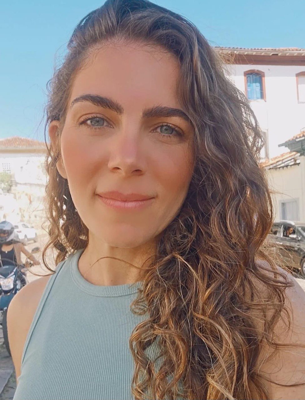 Flávia Maitri sorrindo ao ar livre, com casario histórico de Minas Gerais ao fundo